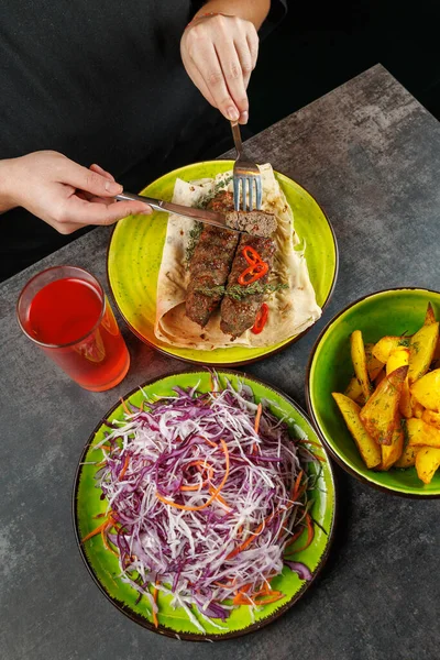 Bir insan dilimlenmiş biberle kaplanmış ızgara et yemeye hazırlanıyor. Yakınlarda da patates kızartması, lahana ve havuç salatası ve bir de içecek vardır..