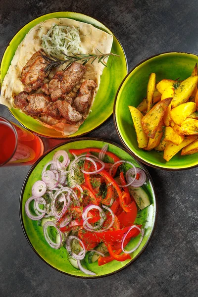 Bir masada kızarmış et ile yassı ekmek, soğanlı taze salata ve renkli kaselerde çıtır patatesler sergilenmektedir. Serinletici bir içecek de mevcut..