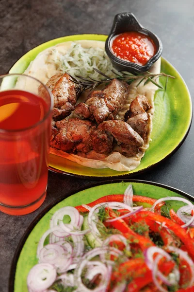 Düz ekmeğin üzerine taze sebzelerle ızgara et konmuş. Renkli bir salata yeşil bir tabakta tabağa eşlik eder. Bir içki yakınlarda oturur, yemek deneyimini tamamlar..