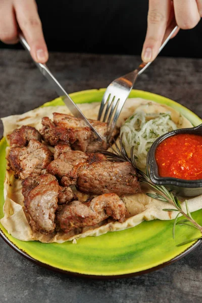 Birisi çatal ve bıçakla kızarmış et parçaları kesiyor. Tabakta sebze salatası ve küçük bir kase kırmızı sos var..