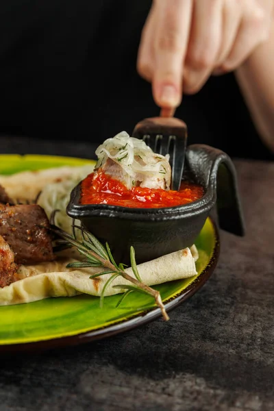 Bir el, canlı yeşil bir tabak üzerinde lezzetli et ve yassı ekmekle birlikte, küçük bir tabakta zengin domates sosu servis etmeye uzanır..