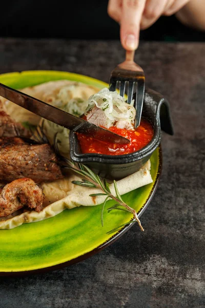 Bir insan küçük dökme bir demir tabaktan et ve sos servis etmek için çatal ve bıçak kullanır. Yemek, canlı yeşil bir tabakta yassı ekmek, sebze ve aromatik bir bitki içeriyor..