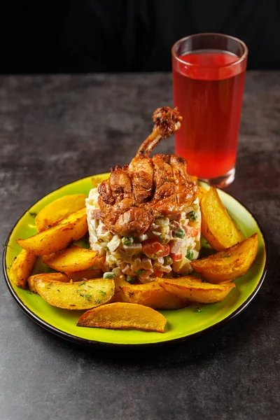 Tabakta sebze salatasının üzerinde kızarmış tavuk ve yanında patates dilimleri yer alıyor. Koyu bir masada, tabağın yanında bir bardak kırmızı içki duruyor..