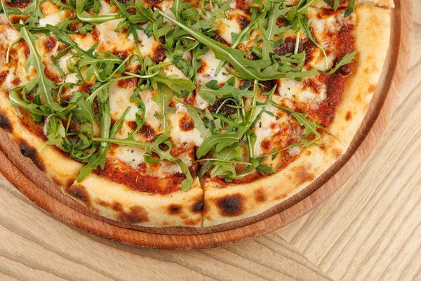 Lezzetli bir pizza, erimiş peynir ve taze roka ile kaplı ahşap bir tahtanın üzerinde oturur. Arka planda basit bir yemek masası görünüyor. Hoş bir ortam yaratıyor..