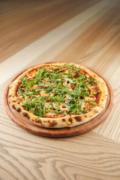 Kıtır kıtırlı yuvarlak bir pizza üzerine taze roka katılmış domates sosu ve peynir katmanı içerir. Tahta bir tahta üzerinde sunuluyor, açık tahta bir yüzeye yerleştiriliyor..