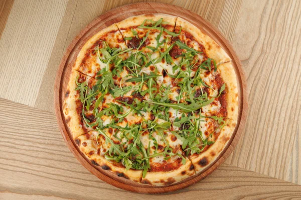 Taze pizza ahşap bir tahtanın üzerinde duruyor. Üstünde bir sürü roka var. Arka plan basit bir tahta masayı gösteriyor. Bu sahne arkadaşlarla yemek yemeye ve eğlenmeye davet ediyor..