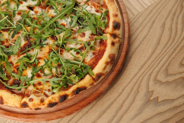 Lezzetli pizza, eritilmiş peynir ve taze roka ile kaplı ahşap bir tabakta duruyor. Sıradan yemek atmosferi bu yemeğin tadını çıkarmak için mükemmel bir ortam hazırlıyor..