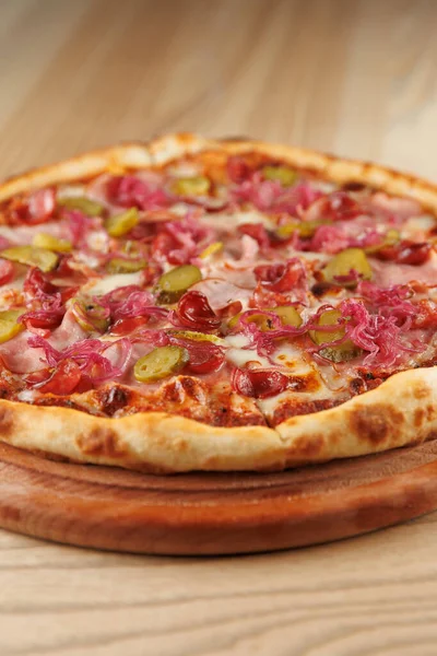 Dilimlenmiş turşu, soğan ve kiraz domatesli taze pişmiş pizza ahşap bir tahtaya yerleştirilir. Kabuk altın gibi görünüyor ve kıtır kıtır, tadını çıkarmaya hazır..