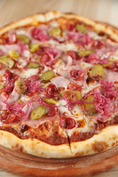 Yuvarlak bir pizza taze pişmiş ve dilimlenmiş jambon, turşu ve kırmızı soğanla kaplıdır. Tabak ahşap bir tabakta sunuluyor, malzemelerin canlı renklerini vurguluyor..