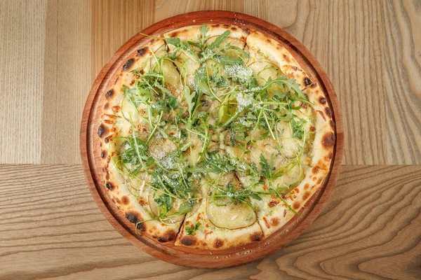 Yuvarlak pizza, taze yeşil malzemeler ve patates dilimleriyle harmanlanmış dokular sergiliyor. Bir restoranın ahşap tahtasında sunuluyor..
