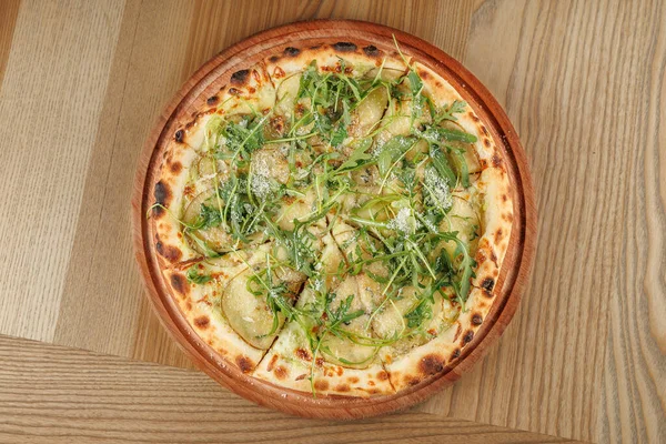 Yeşil otlar ve dilimlenmiş malzemelerle kaplı taze bir pizza ahşap bir tabakta duruyor. Yemek hafif ahşap yüzeyli sıradan bir yemek ortamında servis ediliyor..