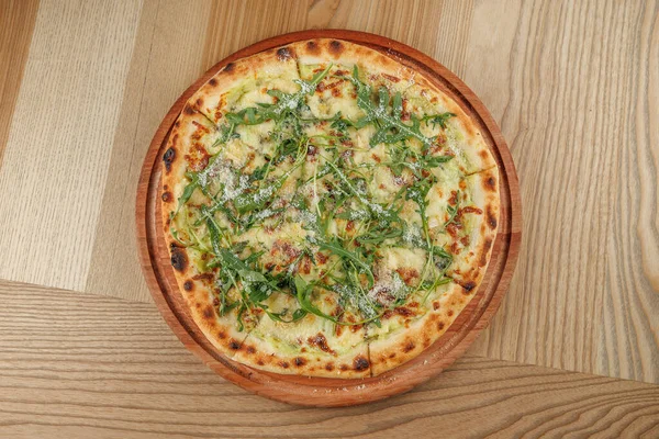 Yuvarlak, ince hamurlu bir pizza yeni pişmiş ve ahşap bir tabakta sunuluyor. İçinde erimiş peynir, taze roka ve kırsal ahşap bir masada parmesan serpiştirilmiş..