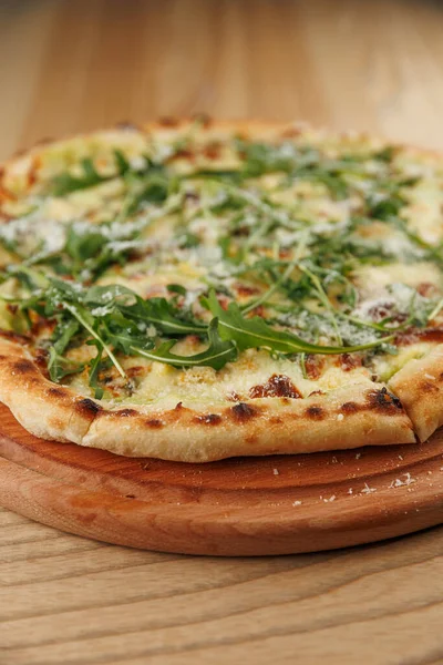 Yeni pişmiş bir pizza ahşap bir tahtanın üzerinde oturur. Pizza bir kat erimiş peynir içeriyor ve üzerinde canlı roka var..