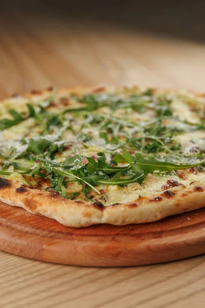 Tahtada pişmiş yuvarlak bir pizza masanın üzerinde oturur. Pizzanın altın hamuru var ve üzeri taze yeşilliklerle kaplı. Sahne sıradan bir yemek atmosferini gösteriyor..