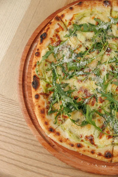 Tahtadan yapılmış bir pizza yuvarlak bir tahtanın üzerinde duruyor. Pizzada taze roka sosu var. Kabuğu altın rengi kahverengi ve çıtır çıtır, erimiş peynir görünür..