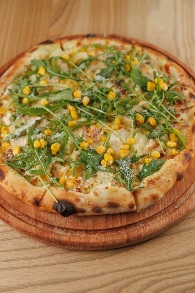 Bir pizza üzerinde erimiş peynir, tatlı mısır ve taze roka ile ince bir kabuk içerir. Tabak, sıradan bir yemek alanında tahta bir tahtanın üzerinde duruyor..