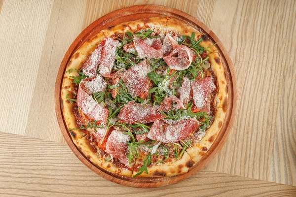 Ahşap tabakta yuvarlak bir pizza, üzerinde pişirilmiş et, taze sebze ve rendelenmiş peynir bulunan bir hamur tabanı içerir. Tabak, gevşek bir lokantada, ahşap bir masaya konur..