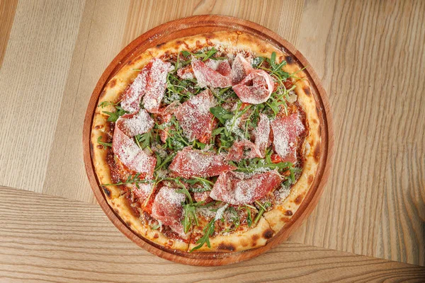 Taze pişmiş pizza, domates sosu, roka ve ahşap bir tabakta peynirle süslenmiş kesilmiş et dilimleriyle kaplı bir kabuk içerir..