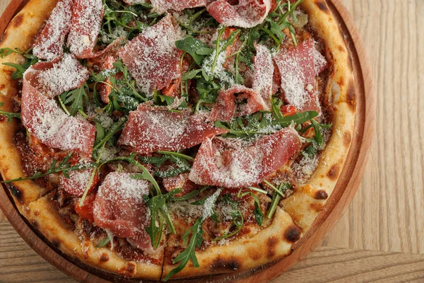 Yuvarlak bir pizza tahta bir tahtanın üzerinde duruyor. Üstlerinde ince et dilimleri ve taze roka var. Pizzanın altın hamuru var ve basit bir ahşap masaya kurulmuş..