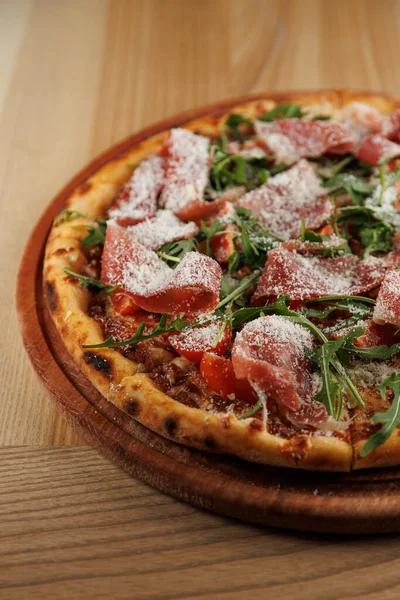 Pizza ahşap bir tahtanın üzerinde oturur. Üstünde jambon, taze roka ve rendelenmiş peynir vardır. Ayar gündelik ve davetkar, paylaşmak için mükemmel..