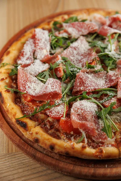 Fırından yeni çıkmış yuvarlak bir pizzada pişmiş et dilimleri, taze roka ve ahşap servis tahtasına serpiştirilmiş peynir bulunur. Pizzada altın hamuru var..