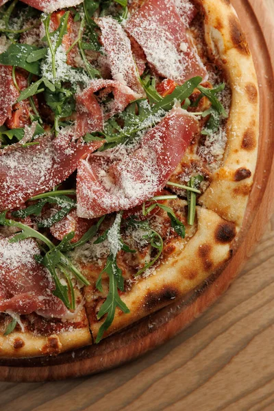 Ahşap bir masa üzerinde ince dilimler halinde jambon, roka ve peynir serpiştirilmiş taze bir pizza sergiliyor. Davetkar atmosfer yemek deneyimini arttırır..