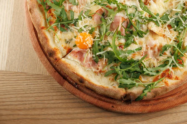 Yeni pişmiş bir pizza ahşap bir tahtanın üzerinde, roka, jambon, peynir ve parlak yumurta sarısı ile kaplıdır. Sahne öğle yemeği sırasında bir kafenin davetkar atmosferini yakalar..