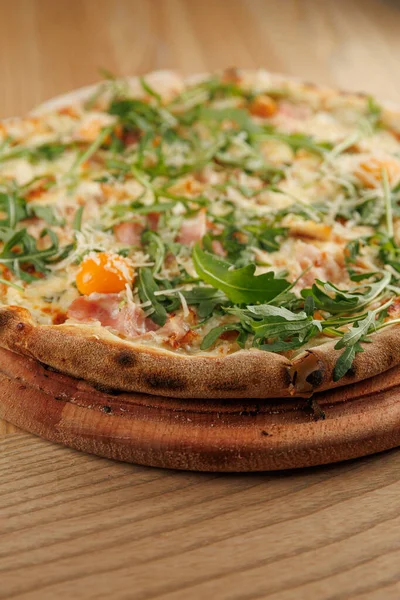 Yeşil, yumurta, peynir ve jambonla kaplı yuvarlak bir pizza sıcak bir restoranda ahşap bir tabakta duruyor. Atmosfer doğal ahşap dekorla davet ediyor..
