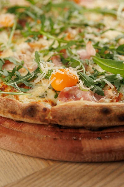 Jambonlu, peynirli, roka aromalı lezzetli bir pizza ve ahşap bir tahtanın üzerinde parlak yumurta sarısı servis edilir. Doğal ışık tabağı aydınlatırken, ortam sıradan..