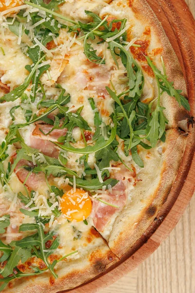 Pizza ahşap bir tabakta, roka, jambon, peynir ve kırık yumurta ile kaplıdır. Sahne, İtalyan mutfağının yer aldığı sıradan bir yemek deneyimini vurguluyor..