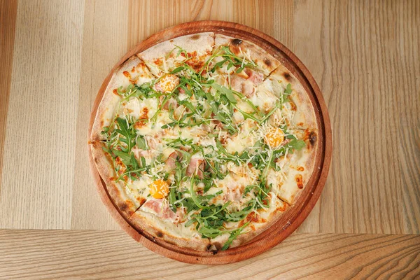 Yeni yapılmış bir pizza, jambon, peynir, roka ve portakal dilimlerini içeren malzemelerle gösterilir. Konforlu bir restoran ortamında ahşap bir tahtada sunuluyor..