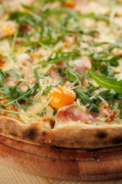 Taze bir pizza üzerine erimiş peynir, roka, jambon parçaları ve ortada çiğ yumurta sarısı. Tabak ahşap bir tahtada servis edilir, keyif verir..