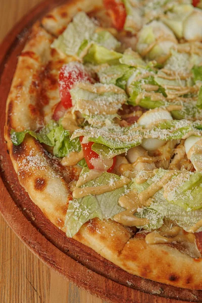 Yuvarlak bir pizza ahşap bir tabakta servis edilir. İçinde salata, domates ve peynir serpintisi gibi malzemeler var. Yemek sıradan bir yemek ortamında sunuluyor..