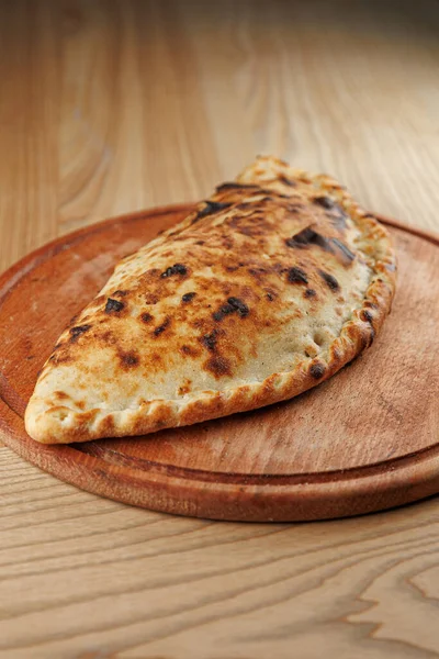 Bir calzone ahşap bir tabakta oturur, altın-kahverengi kabuğunu sergiler ve içini doldurur. Arka plan sıcak atmosferi olan bir köy restoranını işaret ediyor..