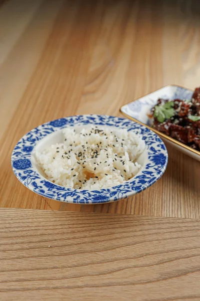 Otlarla serpilmiş bir kase sade pirinç et tabağının yanında oturur. Ahşap masa yemek deneyimine sıcaklık katar..