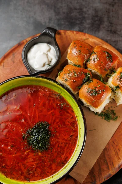 Borş, yeşil bir kaseyi otlarla ve küçük bir tabak ekşi kremayla kaplı yumuşak ruloların yanına doldurur. Tahta tabak bu rahatlatıcı yemeğin sunumunu arttırır..