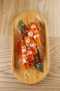 Turuncu balkabağı dilimleri ahşap bir kasede yeşil yaprakların üzerine yerleştirilir. Üzerinde rendelenmiş hindistan cevizi ve sos var. Renkli bir düzenleme sergiliyor..