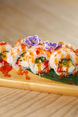 Suşi ruloları ahşap bir tabakta güzelce düzenlenir. Her rulo taze malzemelerle doludur ve parlak sos ve yenilebilir çiçeklerle kaplıdır..