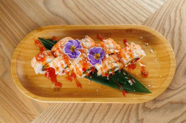 Suşi dürümleri ahşap bir tabakta, sos ve çiçeklerle süslenir, bir restoranda renkli ve çekici bir sunum sergilenir..