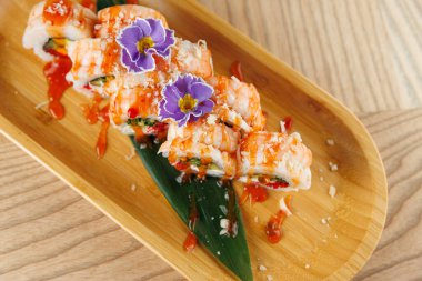 Karides ve sosla kaplı suşi ruloları ahşap bir tabakta servis edilir. Yenilebilir çiçekler yemeğe renk katar. Restoran ortamında eğleniyoruz..