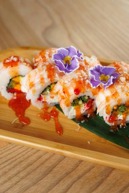 Suşi ruloları ahşap bir tabakta düzenlenir. Ekmeğin üzerinde sos ve yenilebilir çiçeklerle süslenmiş canlı dolgular var. Ayarlar bir Japon restoranında..