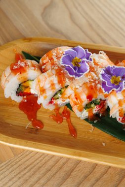 Suşi ruloları karides, sebze ve yenilebilir çiçeklerle kaplı ahşap bir tabakta sunulur. Bu yemek sıradan bir yemek deneyimi ya da özel bir durum için uygundur..