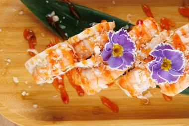 Karidesle dolu suşi ruloları ahşap bir tahta üzerinde düzenlenir. Parlak renkli çiçekler en üste konur, tabağın sunumunu güçlendirir.