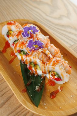 Ahşap bir tepside karides, sos ve yenilebilir çiçeklerle süslenmiş güzel bir suşi rulosu sunulur. Tabakta canlı renkler ve taze malzemeler paylaşım için mükemmel..