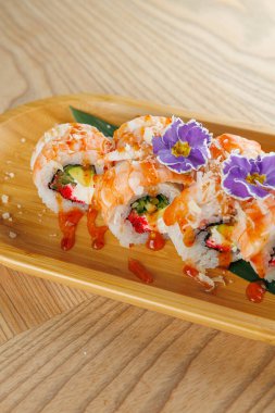 Taze suşi ruloları karides, sos ve çiçeklerle kaplı ahşap bir tabakta servis edilir. Davetkar atmosfer yemek deneyimini arttırıyor, suşi severler için mükemmel..