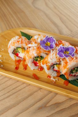 Suşi ruloları ahşap bir tepside canlı renkler ve bir sos yağmuru ile düzenlenir. Taze sebze ve çiçekler bu mutfak yaratımına dekoratif bir dokunuş ekler..