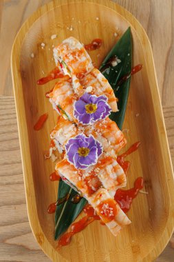 Karidesle doldurulmuş suşi ruloları ve ahşap bir tabakta sosla servis edilir. Yemek, yenilebilir çiçek ve yapraklarla süslenerek, yiyeceklerin sunumunu vurguluyor..