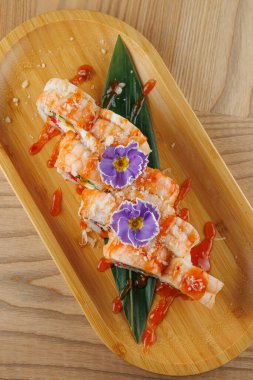 Suşi rulosu ahşap bir tabakta sosla süslenmiş ve yenilebilir çiçeklerle süslenmiştir. Bu yemek bir yemek ortamında lezzet ve sunum karışımı sergiler..