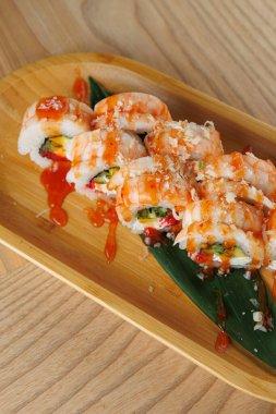 Suşi ruloları ahşap bir tabakta, altında yeşil bir yaprak ile güzelce düzenlenir. Ekmekler sosla kaplanmış ve süslenmiş. Dekor, Japon restoranlarının tipik hafif ahşap yüzeylerini gösteriyor..