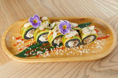 Suşi ruloları ahşap bir tabakta güzelce düzenlenir. Üstlerinde dilimlenmiş badem ve yenilebilir çiçekler var. Renkli bir sunum masada keyif almaya ve paylaşmaya davet eder.